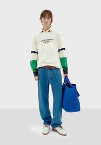 Camisa blanca de manga larga con mangas a rayas verdes y azules, jeans azules, zapatos blancos y una bolsa de mano azul. El texto en la camisa dice "AQUÍ VIENE EL SOL."