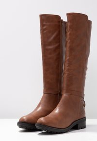 Bottes en cuir marron jusqu'au genou avec fermeture éclair latérale, talon bas et sangle décorative avec anneau en métal sur surface blanche.