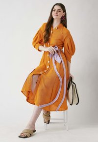 Oranssi nappikauluspaita mekko, jossa on puffihihat ja laventelinvärinen vyö. Siinä on halkio ja teksturoitua kangasta. Yhdistetty beigeihin sandaaleihin ja laukkuun.