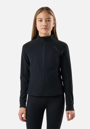 Ung person stående iført en sort lynlåstrøje og sorte bukser, vendt mod kameraet foran en ensartet lys baggrund.