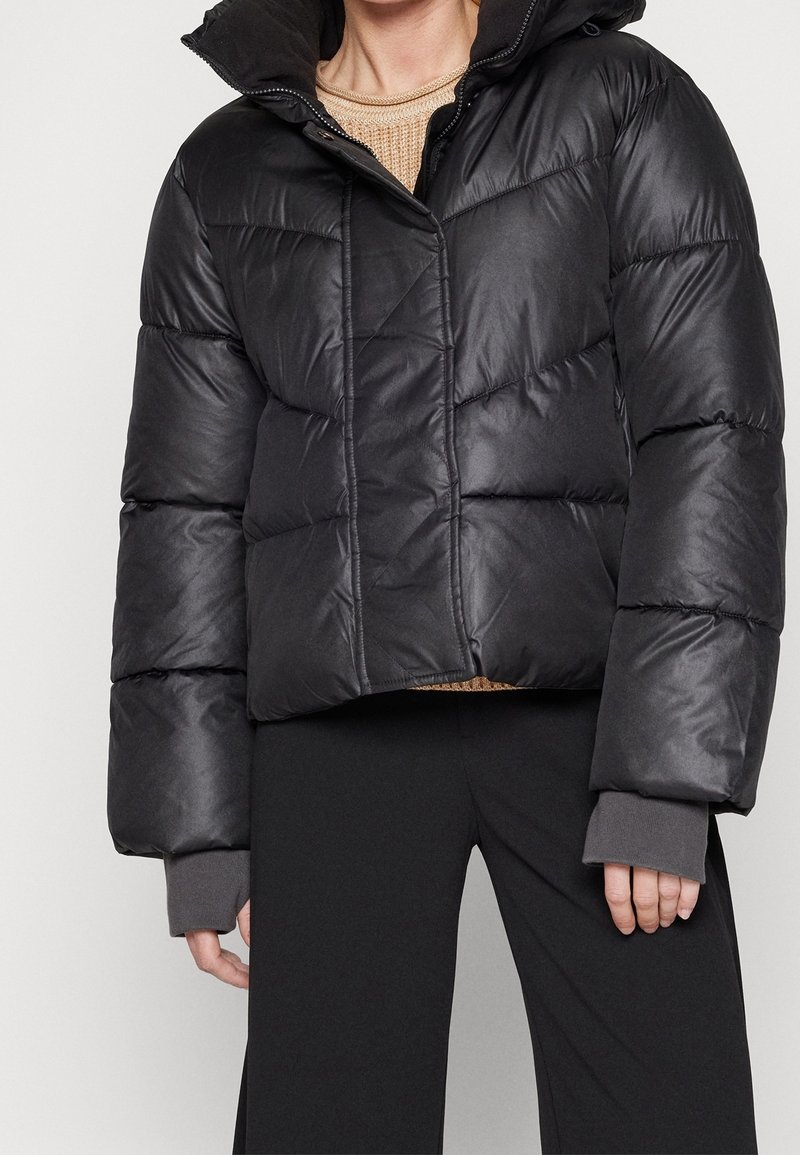 Personne portant une veste matelassée noire avec un col montant sur un pull en maille beige et un pantalon noir, debout devant un arrière-plan uni.