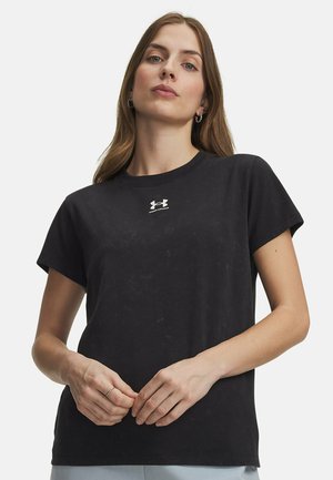 Vrouw met lang bruin haar, die een zwart Under Armour T-shirt draagt, lichte broek en zilveren creooloorbellen, staand tegen een witte achtergrond.
