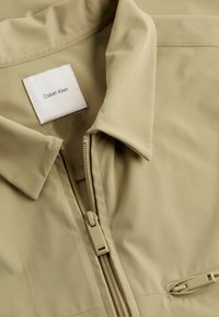 Beige Zip-Jacke mit Kragen, aus glattem Material gefertigt. Verfügt über einen frontalen Reißverschluss und eine kleine Seitentasche mit verborgenem Reißverschluss.