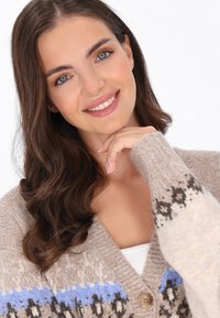 Beige kötött kardigán V-nyakkal, kék és fekete geometrikus mintákkal a ujjakon, nagy gombbal és bordás mandzsettákkal befejezve.