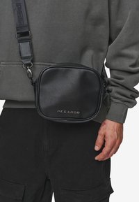 Sort crossbody taske med en glat syntetisk tekstur, rektangulær form, justerbar skulderrem og præget "PEGADOR" logo foran.