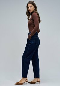 Top marrón de manga larga combinado con pantalones de denim azul oscuro de cintura alta, con pierna recta y sutiles detalles desgastados, y zapatos de tacón marrones.