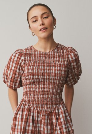 Mladá žena s vlasy staženými dozadu nosí šaty s rezavě bílým kostkováním, nabíranými rukávy a nařaseným živůtkem, stojící před jednobarevným pozadím.