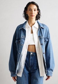 Ljusblå denimjacka med stora framfickor och bruna knappar, bärs över en vit skjorta med knappar och matchande höga jeans.