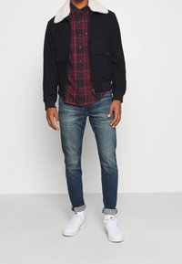 Giacca nera con colletto in pelliccia bianca, camicia a quadri rossi e blu navy, jeans in denim blu e sneaker bianche. Presenta chiusure con zip e bottone.