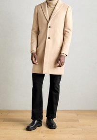 Abrigo de lana beige con solapa estructurada, diseño de un solo botón, dos botones y bolsillos laterales inclinados. Combinado con pantalones negros y botas.