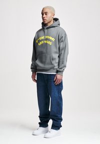 Sudadera grigia con cappuccio con testo giallo a bolle "F*** YOUNG BAD VIBES," abbinata a jeans blu e sneaker bianche, dotata di tasca frontale.