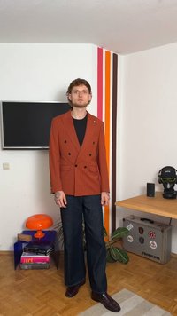 Blazer arancione a righe con doppiopetto e bottoni neri, abbinato a pantaloni in denim scuro e scarpe marroni, su uno sfondo di muro colorato.