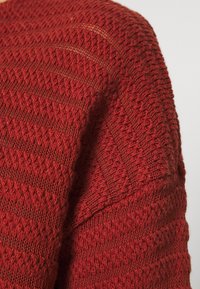Maglione rosso lavorato a maglia con un design testurizzato e a motivo. Presenta un mix di punti a maglia lisci e a coste, evidenziando un aspetto caldo e accogliente.