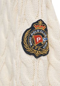 Sweter w kolorze kremowym, wykonany na drutach, z wyraźną czarną i złotą haftowaną naszywką z logo, zdobioną detalem korony i motywami polo.