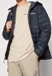 Kurtka puchowa w ciemnym granatowym kolorze z pikowanym wzorem, zamkiem błyskawicznym z przodu, kapturem i logo Columbia. Nosi się ją na jasnobeżowej koszulce i beżowych spodniach.