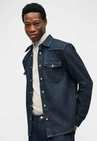 Νεαρός άνδρας που φορά μπλε σκούρο τζιν μπουφάν και τζιν παντελόνι, με μπεζ πουλόβερ με φερμουάρ, ποζάρει με τα χέρια πίσω από την πλάτη σε απλό φόντο.