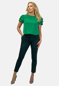 Zielona bluzka z falbanami na rękawach, w połączeniu z ciemnozielonymi spodniami typu slim-fit i wysokimi obcasami w wężowy wzór. Gładki materiał i dopasowany krój.