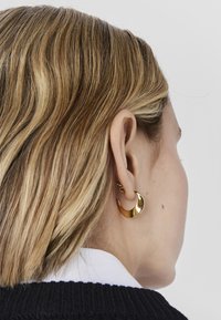 Donna con capelli corti e mèches indossa un orecchino a cerchio a forma di mezzaluna dorato lucido e un maglione nero sopra una camicia bianca.