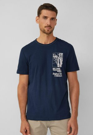 Man in een marineblauwe t-shirt met een witte boomafbeelding en de tekst "Omarm het Onbekende", gecombineerd met beige broek, staande tegen een grijze achtergrond.