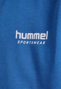 Blå bomull T-shirt med "hummel"-logotyp i vitt, med "SPORTSWEAR" nedanför. Tyget har en slät textur och är något elastiskt.