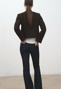 Giacca cropped marrone dalla texture elegante con maniche lunghe, abbinata a jeans svasati scuri, vista da dietro su uno sfondo chiaro.