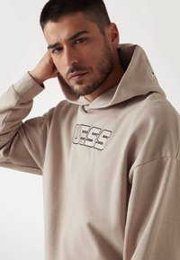 Beige kapucnis pulóver, melyen a "GUESS" szó fekete hímzéssel található. Lazán illeszkedő fazonnal és puha, sima textúrával rendelkezik.