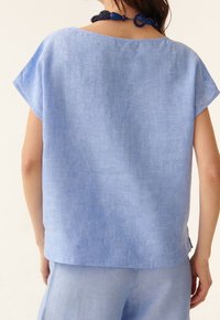 Blusa de lino ligera y holgada de color azul claro con mangas cortas y escote redondeado. Presenta una textura suave y sutiles variaciones en la tela.