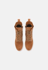 Bottines en daim marron avec des lacets beige, bout arrondi, tirette en boucle et semelle en caoutchouc blanc. Intérieur doublé de matériau foncé.