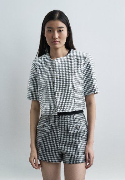 Femme portant une veste à manches courtes en tweed noir et blanc et un short taille haute assorti, debout devant un fond clair uni.