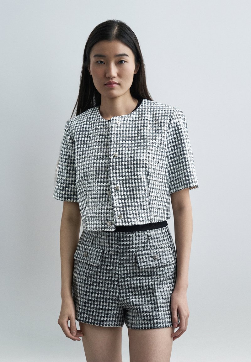 Femme portant une veste à manches courtes en tweed noir et blanc et un short taille haute assorti, debout devant un fond clair uni.