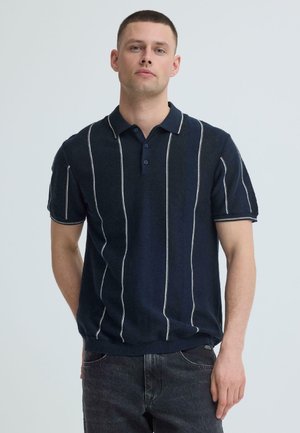 Man wearing navy blue short-sleeve polo shirt with vertical white stripes and dark jeans, standing against a plain light background.