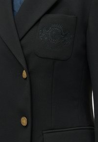 Črn blazer z zlatimi gumbi, vezeninasti grb z levom, valovi in krono nad trakom z napisom "Hilfiger" na žepu prsih.