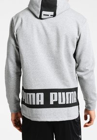 Grå huvtröja med en svart panel på huven. Har en stor svart "PUMA"-logotyp på baksidan och ribbade muddar för en tight passform.