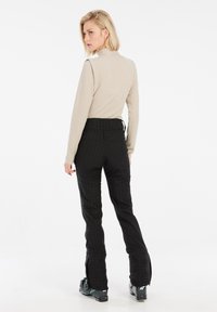 Hellbeige gestreiftes Langarmtop, kombiniert mit schwarzen strukturierten Skihosen mit seitlichen Reißverschlüssen und schwarzen Skistiefeln, von hinten gezeigt.