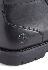 Gros plan d’une botte en cuir noir montrant un logo arbre embossé et le texte « Imperméable » sur le côté au-dessus d’une semelle épaisse en caoutchouc rainurée.