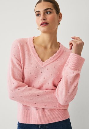 Jonge vrouw met bruin haar in een staart die een roze V-hals trui draagt met kleine opengewerkte patronen en een blauwe spijkerbroek, poserend met één arm omhoog.