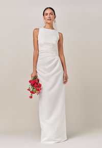 Vit sidenklänning med figurnära siluett, ärmlös design och draperade detaljer på sidan. Håller en bukett med röda blommor. Enkel, elegant stil.