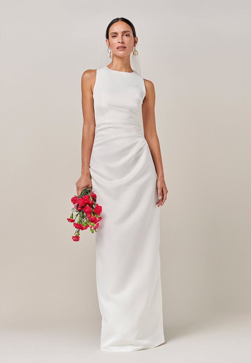 Vit sidenklänning med figurnära siluett, ärmlös design och draperade detaljer på sidan. Håller en bukett med röda blommor. Enkel, elegant stil.