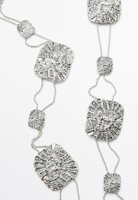 Halsband i silverfärg med texturerade fyrkantiga och rektangulära metallringar, sammanlänkade med snodda kedjetrådar.