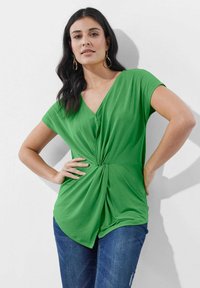 Grünes T-Shirt mit V-Ausschnitt und kurzen Ärmeln, mit einem gedrehten Frontdetail. Kombiniert mit blauen Jeans. Glatte Textur, lässige Passform.