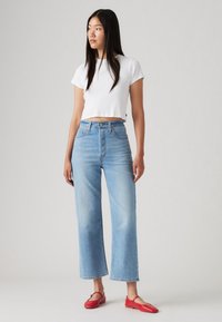 Jauna sieviete, tērpusies baltā īsā krekla, augstās jostasvietas gaiši zilos džinsos un sarkanos baleta zābakos, stāv pret vienkrāsainu fonu.