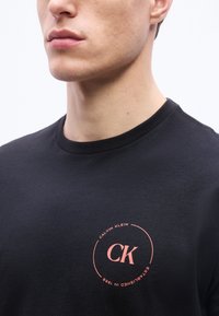 Νεαρός άνδρας που φοράει μαύρο t-shirt Calvin Klein με μικρό ροζ κυκλικό λογότυπο στο στήθος.