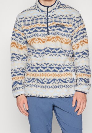 Man met een fleece trui met een patroon van blauwe en oranje geometrische strepen, gecombineerd met een blauwe broek.