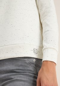 Gros plan sur une main reposant près de la hanche, portant un sweat-shirt blanc à pois portant l'inscription "Vue sur le lac" et un jean en denim gris.