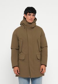 Oliven grøn parka med høj krave, store frontlommer og lynlåslukning. Har hætte og struktureret stof.