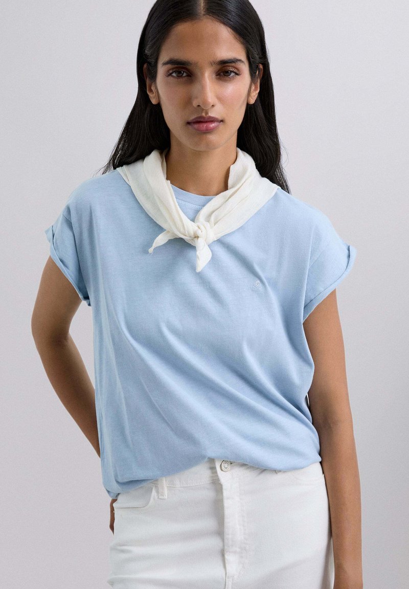 Camiseta de manga corta azul claro con puños remangados, combinada con pantalones blancos. Una bufanda blanca está atada al cuello. Textura suave.