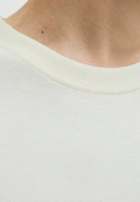 Prim-plan al gâtului și pectoralului unui bărbat care poartă un tricou simplu de culoare albă-crem, cu guler rotund și model cu nervuri.