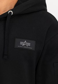 Sudadera negra de tela suave que presenta un parche frontal con el texto "Alpha Industries", cordones ajustables y detalle de bolsillo tipo canguro.