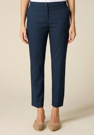 Donna che indossa pantaloni su misura blu navy alla caviglia con bottone e zip davanti, abbinati a scarpe basse beige intrecciate.