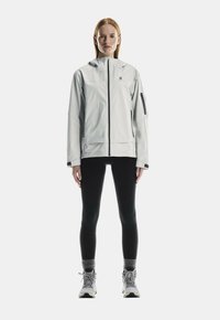 Ljusgrå vattentät jacka med huva, svart dragkedja och sidoficka. Bärs med svarta tights och grå vandringsskor. Minimalistisk design.